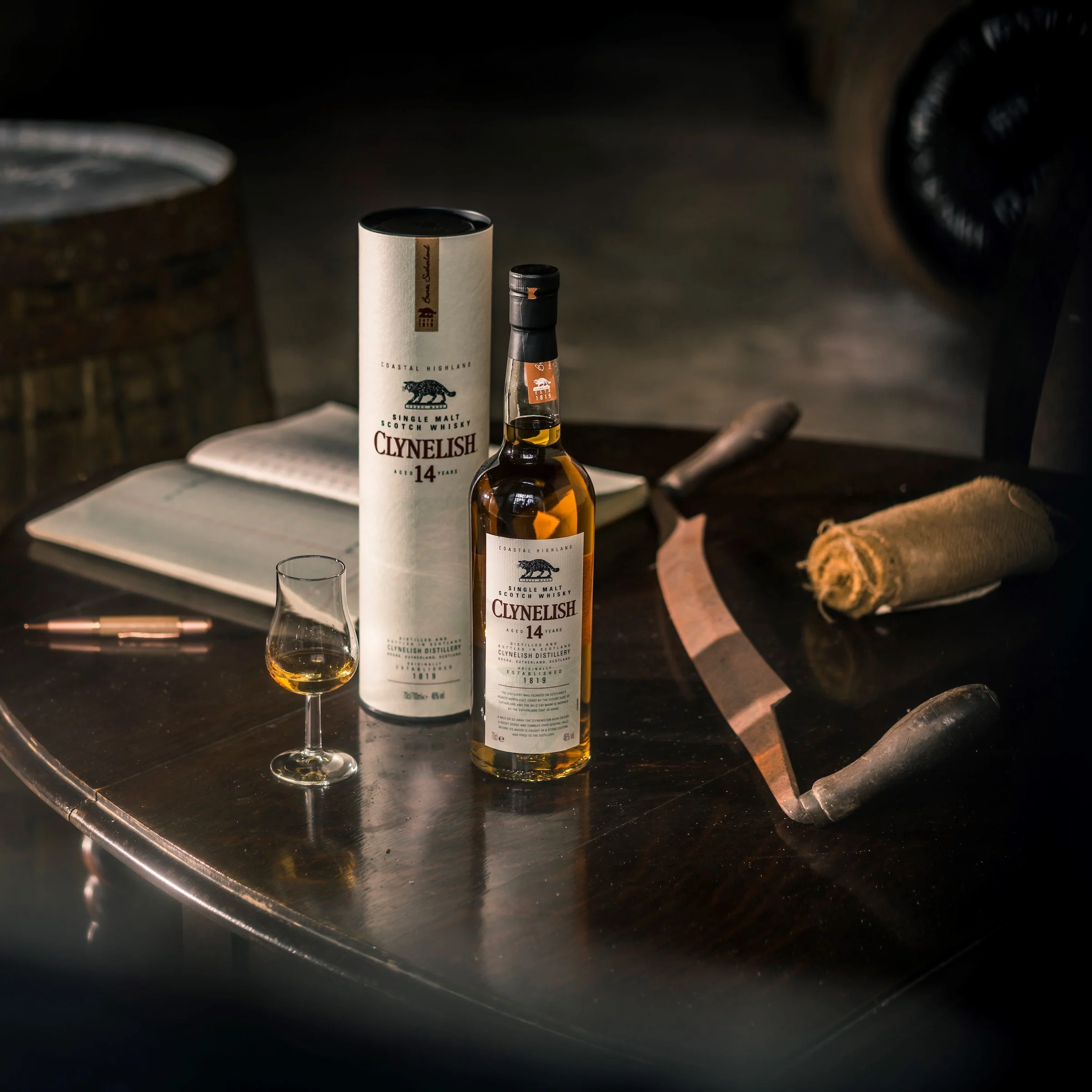 A bottle of Clynelish whisky sits on a circular wooden table, with its outer tube standing behind it. Also on the table is a dram glass containing whisky, a pen, notebook and a rolled-up napkin. Behind the table is a wooden barrel