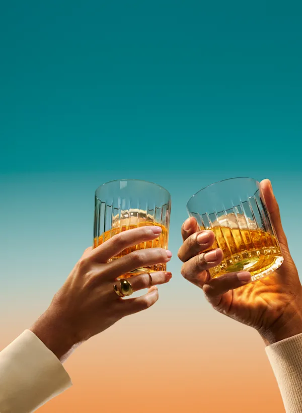 Discover Two hands hold a rocks glass each and bring them together in a ‘cheers’ motion. The glasses contain a large ice cube and whisky.