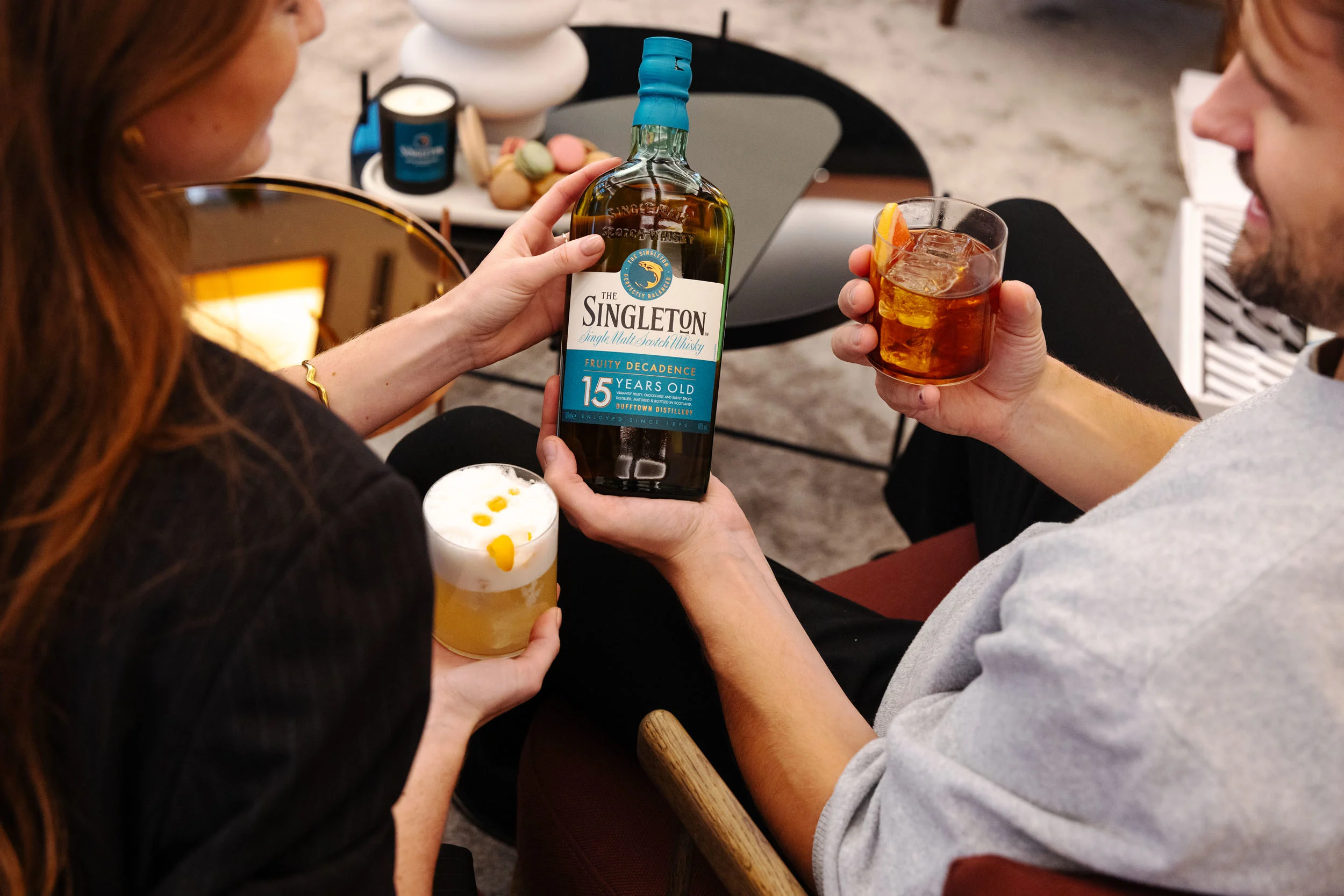 A bottle of Johnnie Walker black label on a yellow table lying down, next to it is string, scissors and a tall long whisky cocktail.