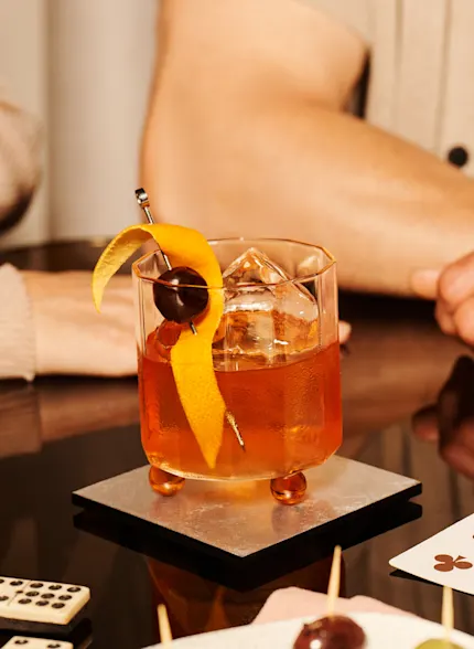 An Old Fashioned cocktail sits on a table by canapes, dominoes and cards