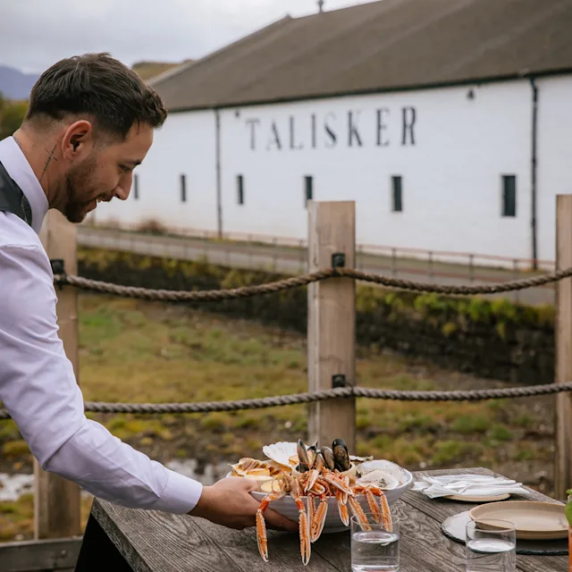 A waiter places a tray of langoustines onto a table which is outside overlooking a Scottish loch. In front of him is the Talisker distillery.