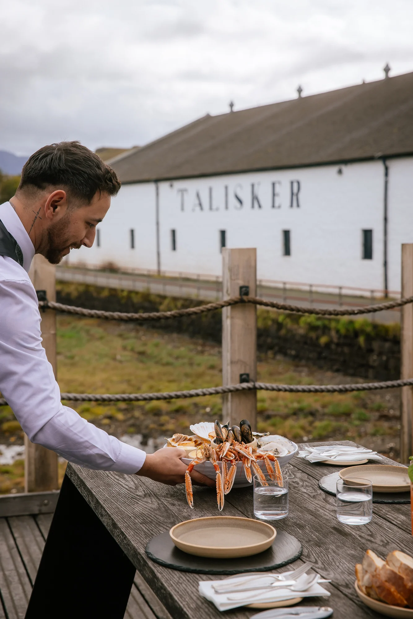 A waiter places a tray of langoustines onto a table which is outside overlooking a Scottish loch. In front of him is the Talisker distillery.
