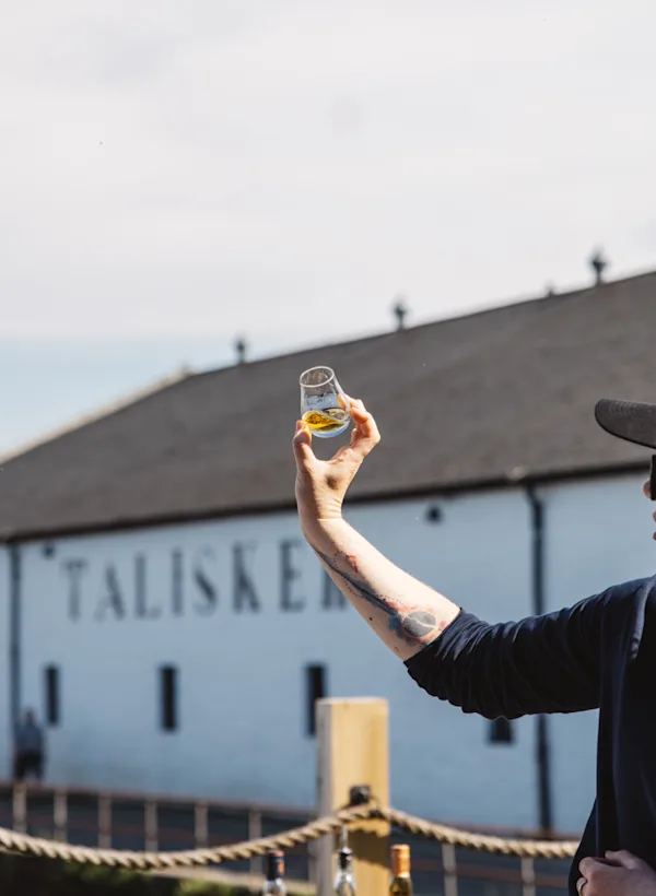 A man stood outside the distillery looking at a glass of talisker against the sky