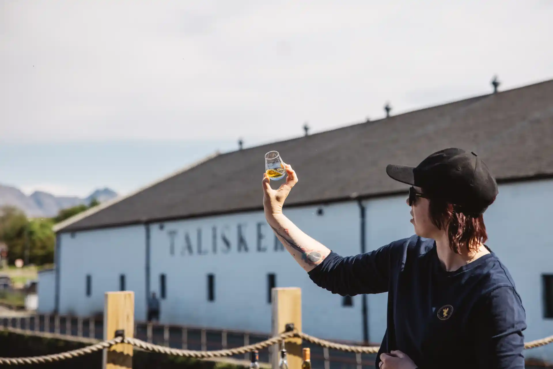 A man stood outside the distillery looking at a glass of talisker against the sky