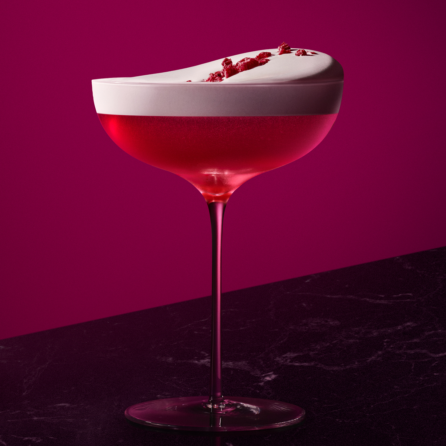 A vibrant red cocktail in a coupe glass, garnished with frothy foam on top, raspberries, and a sprig of mint. The drink is set against a deep magenta background on a dark surface.