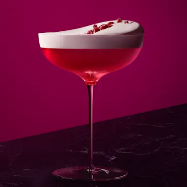 A vibrant red cocktail in a coupe glass, garnished with frothy foam on top, raspberries, and a sprig of mint. The drink is set against a deep magenta background on a dark surface.