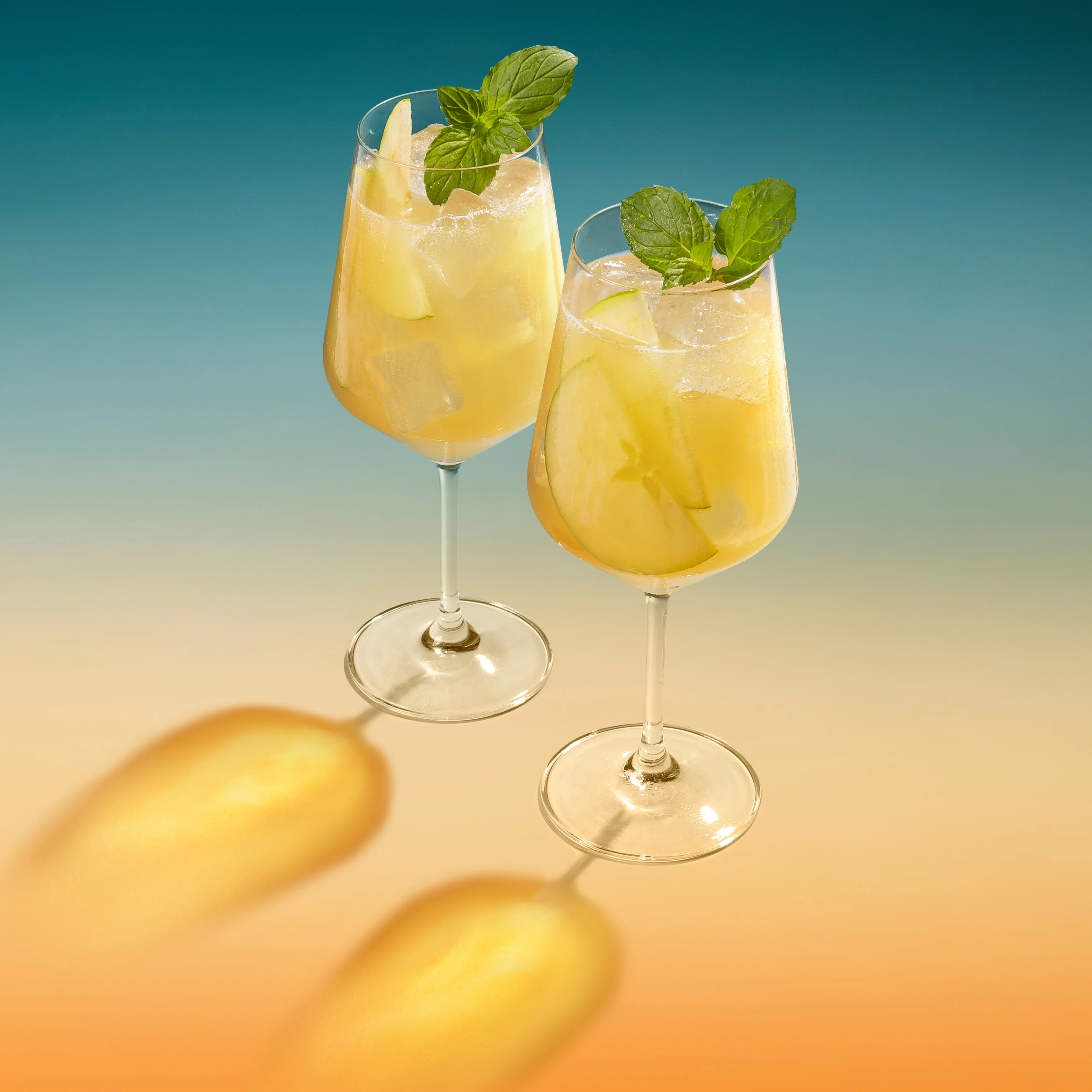 Two elegant glasses filled with a refreshing The Singleton Apple Spritz cocktail, garnished with mint leaves, set against a gradient background of blue and orange.