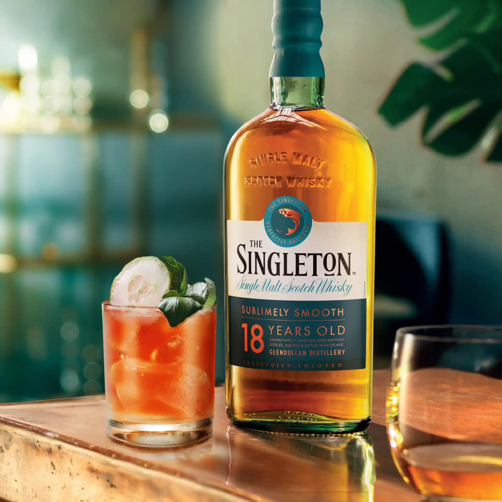 Eine Gruppe von Personen hält über einem Tisch mit brennenden Kerzen Steingläser mit Whisky-Cocktails hoch. Eine Person hält eine Flasche The Singleton 12-year-old Single Malt Scotch Whisky mit dem Etikett in die Kamera.