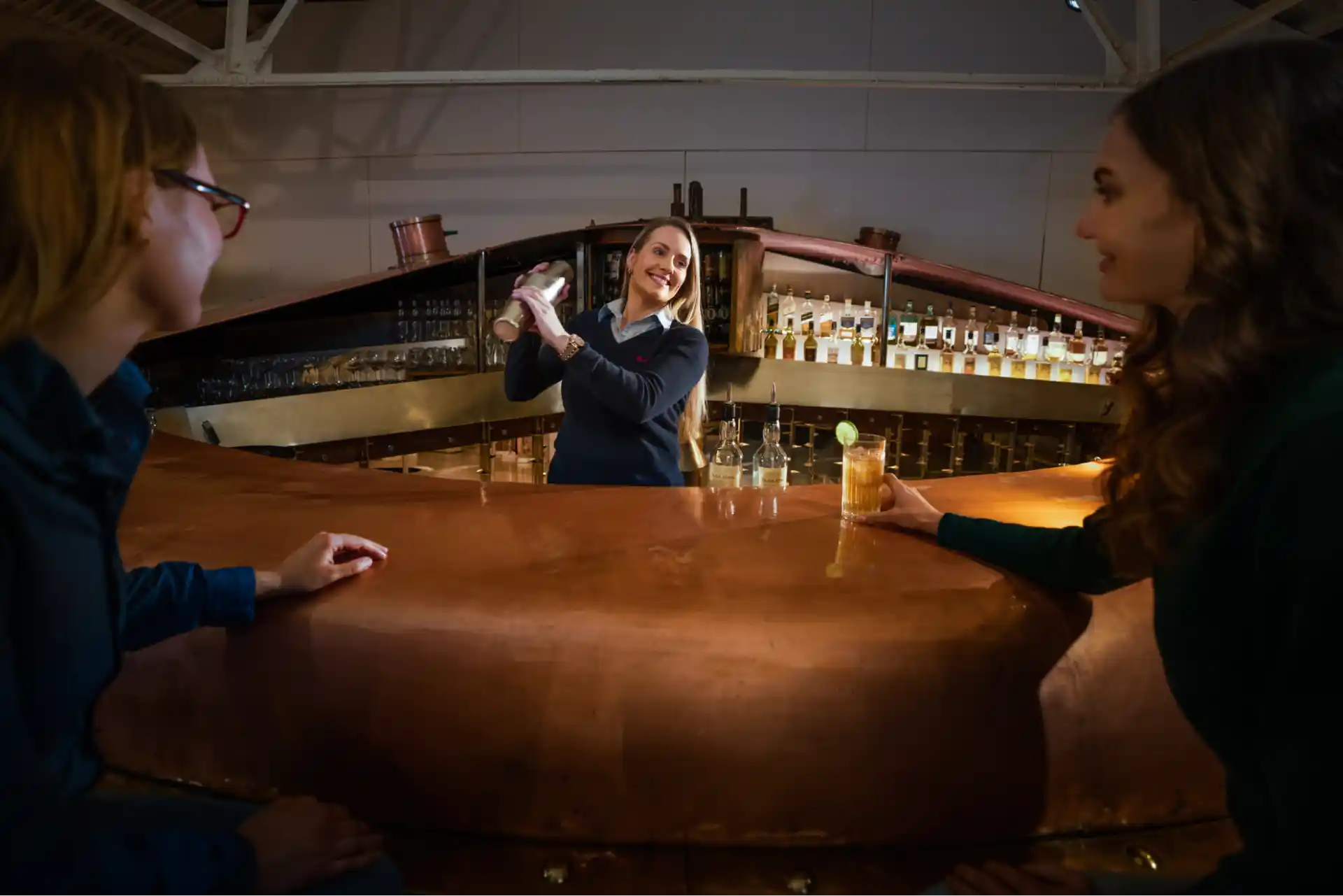 A bartender stands behind a bar, shaking a cocktail shaker. Two guests, one who holds a cocktail in a highball glass and one who waits for the bartender to make the next cocktail.