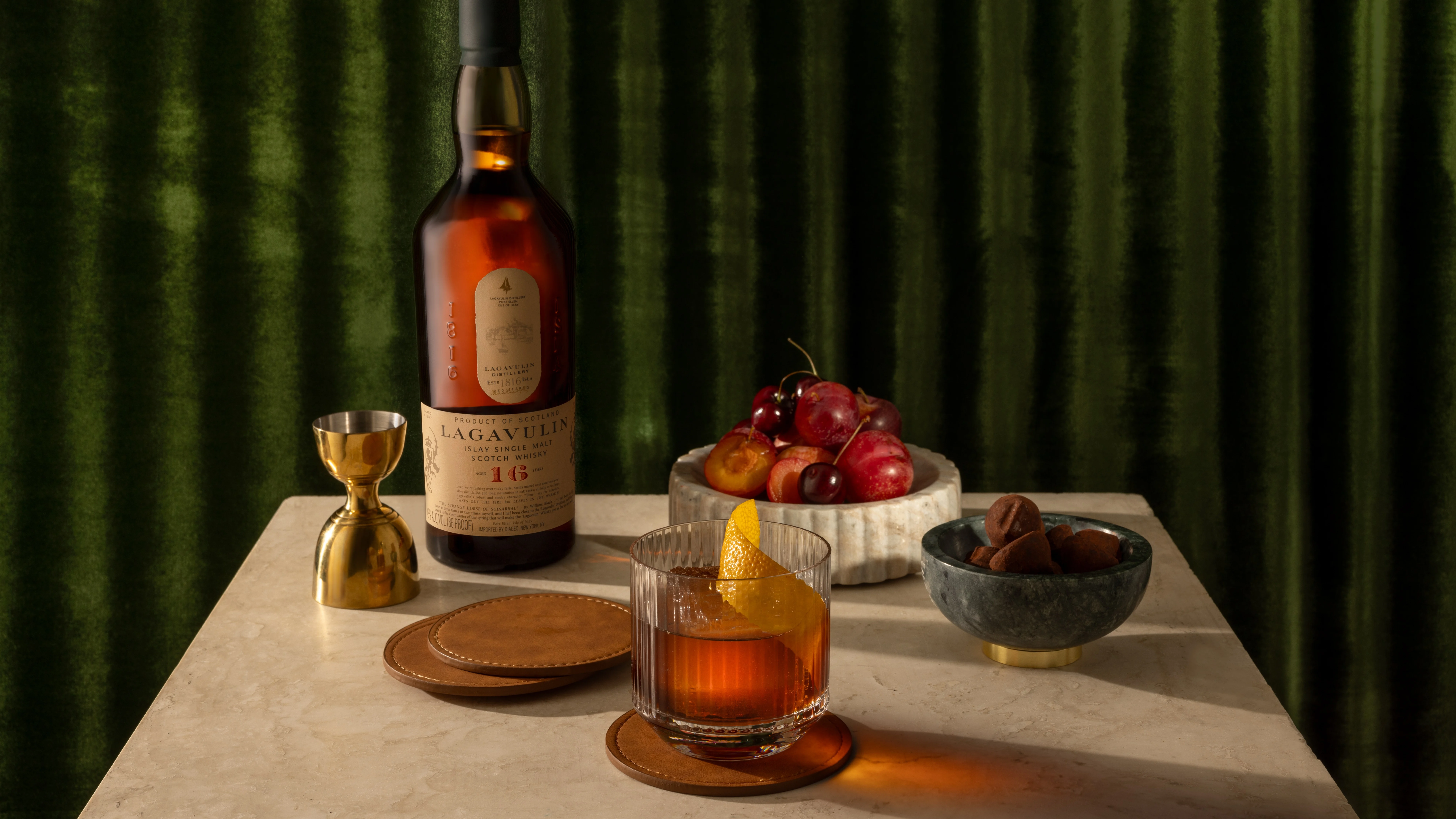 A cocktail next to a bottle of Lagavulin 16 and bowls of fruit
