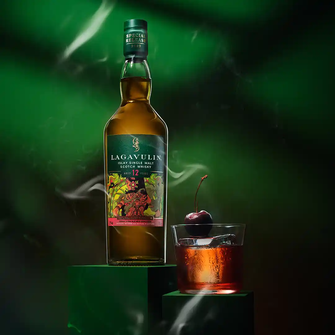 A bottle of Lagavulin 12 year old whisky sits on a green plinth next to a rocks glass holding whisky and a cherry