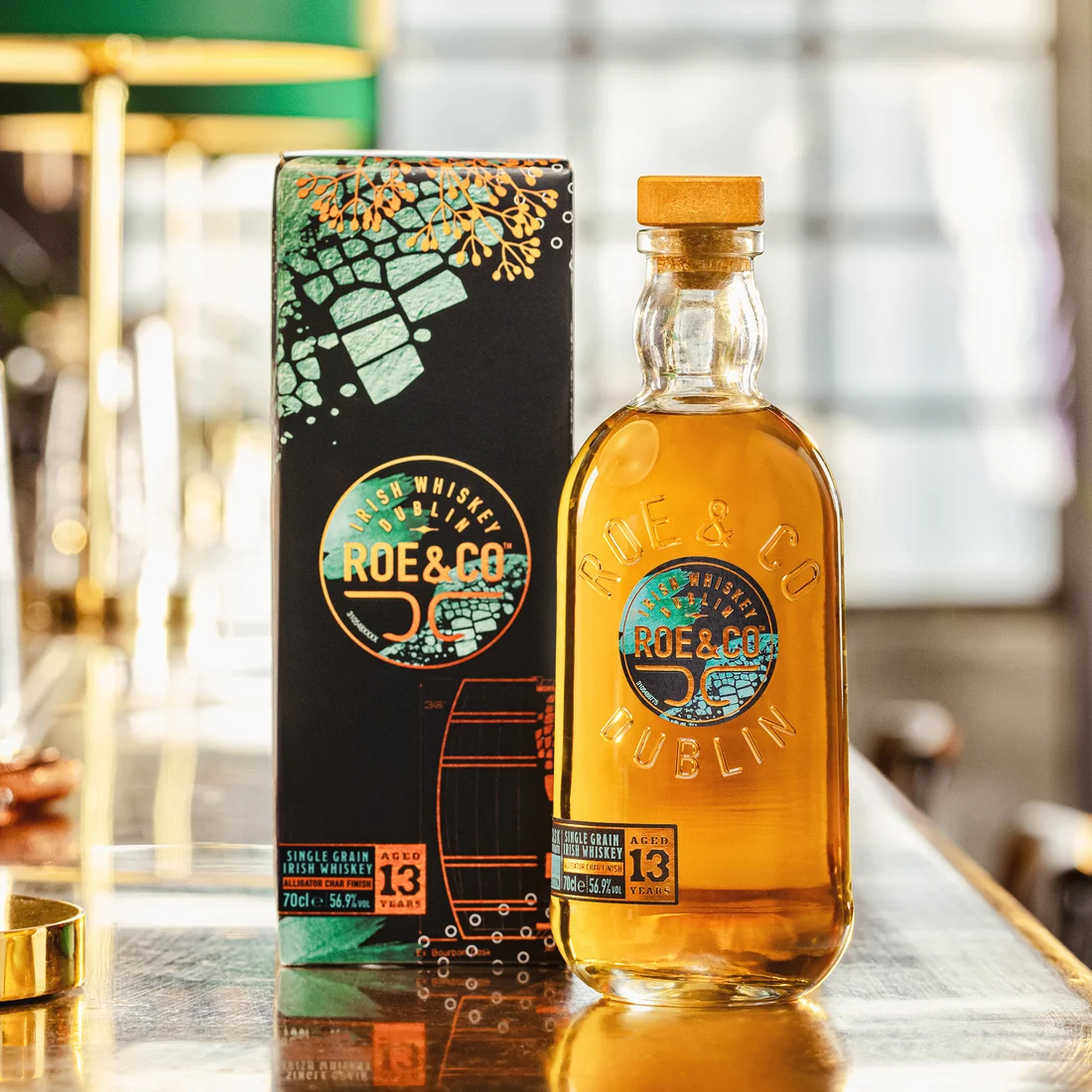 Die Flasche Roe & Co 13-Year-Old Cask Strength Irish Whiskey steht neben der Verpackungsschachtel auf einer Barfläche mit warmer Beleuchtung.