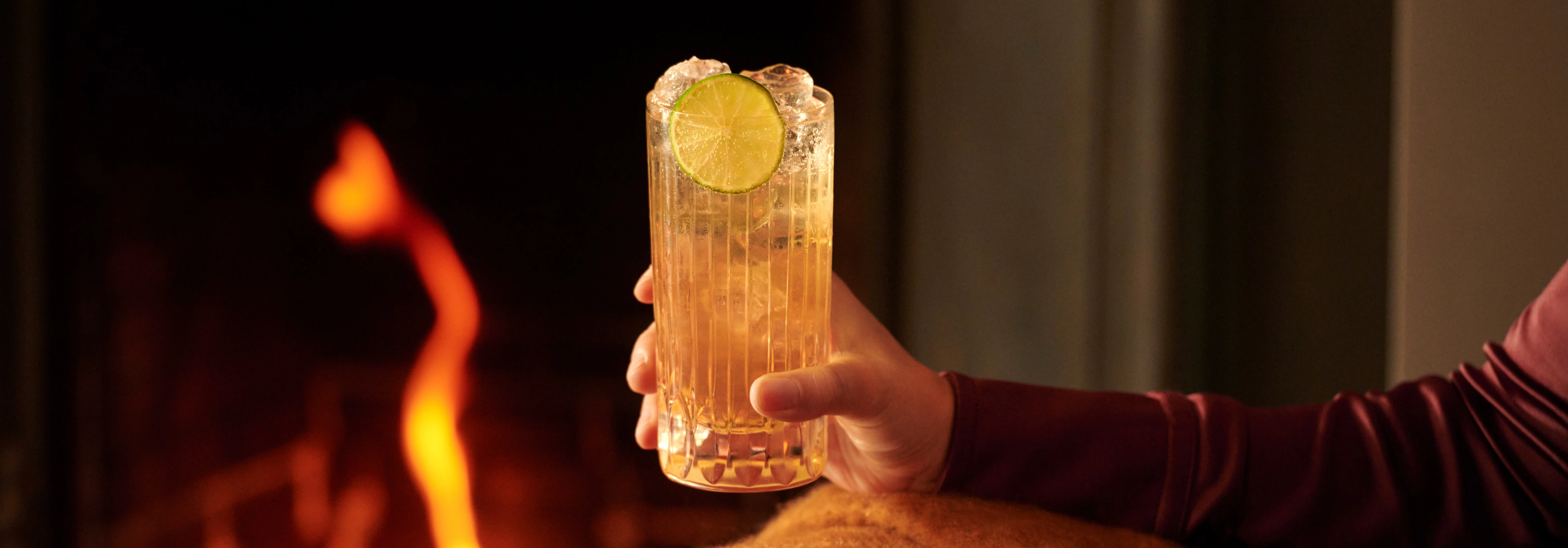 A person sits in an armchair by a fire, holding a whisky highball cocktail