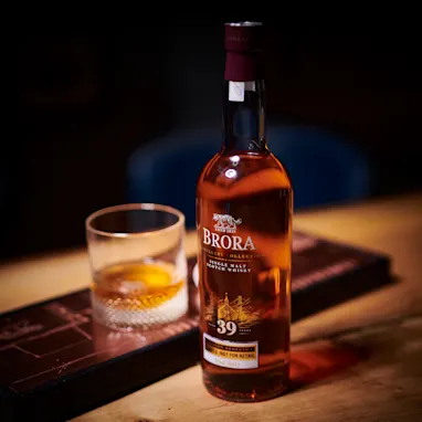 Een fles Brora 39 jaar oude single malt Schotse whisky staat op een houten tafel onder een spot. Ernaast staat een glas met ijs en whisky.