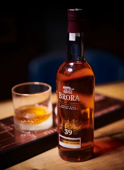 A bottle of Brora 39-year-old single malt Scotch whisky sits on a wooden table under a spotlight. Next to it sits a rocks glass with whisky.