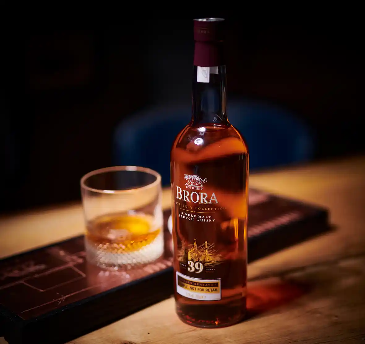 A bottle of Brora 39-year-old single malt Scotch whisky sits on a wooden table under a spotlight. Next to it sits a rocks glass with whisky.