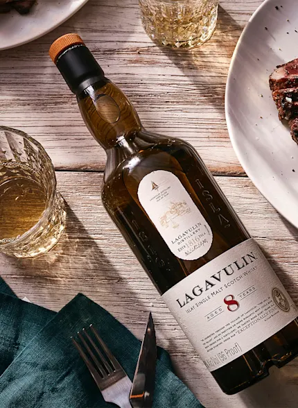Eine Flasche Lagavulin 8 Jahre alter Whisky liegt auf dem Rücken auf einem Holztisch, umgeben von Tellern mit Speisen und Steingläsern mit Whisky