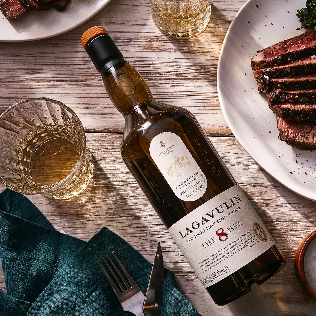 A bottle of Lagavulin 8 year old whisky lies on its back on a wooden table, surrounded by plates of food and rocks glasses containing whisky