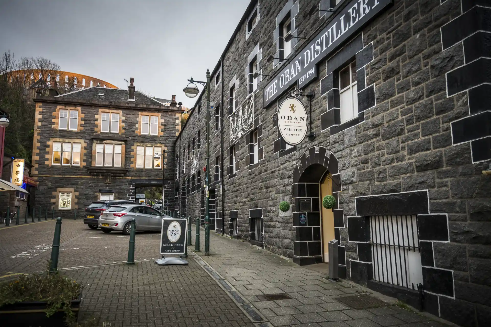 Die graue, schwarz-weiße Backsteinfassade der Oban Distillery ist malerisch, und an der Wand ist ein Schild mit der Aufschrift "Oban Distillery, Visitor Centre" angebracht. Die Destillerie liegt an einer mit Ziegeln gepflasterten Straße.