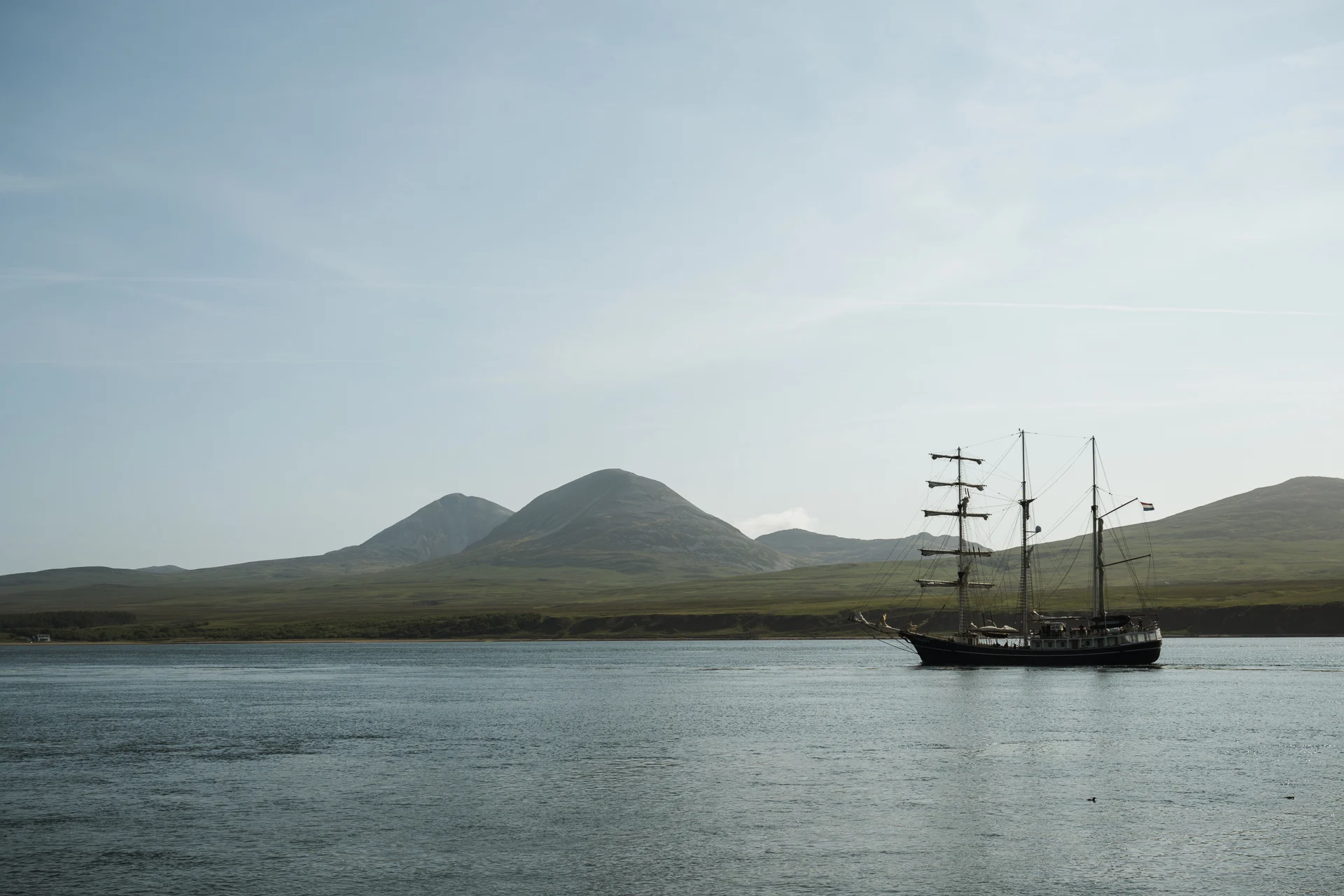 A tall ship with dark sails anchored in the calm waters of a Scottish sea loch on the Isle of Islay, with rolling green hills in the background under a pale sky