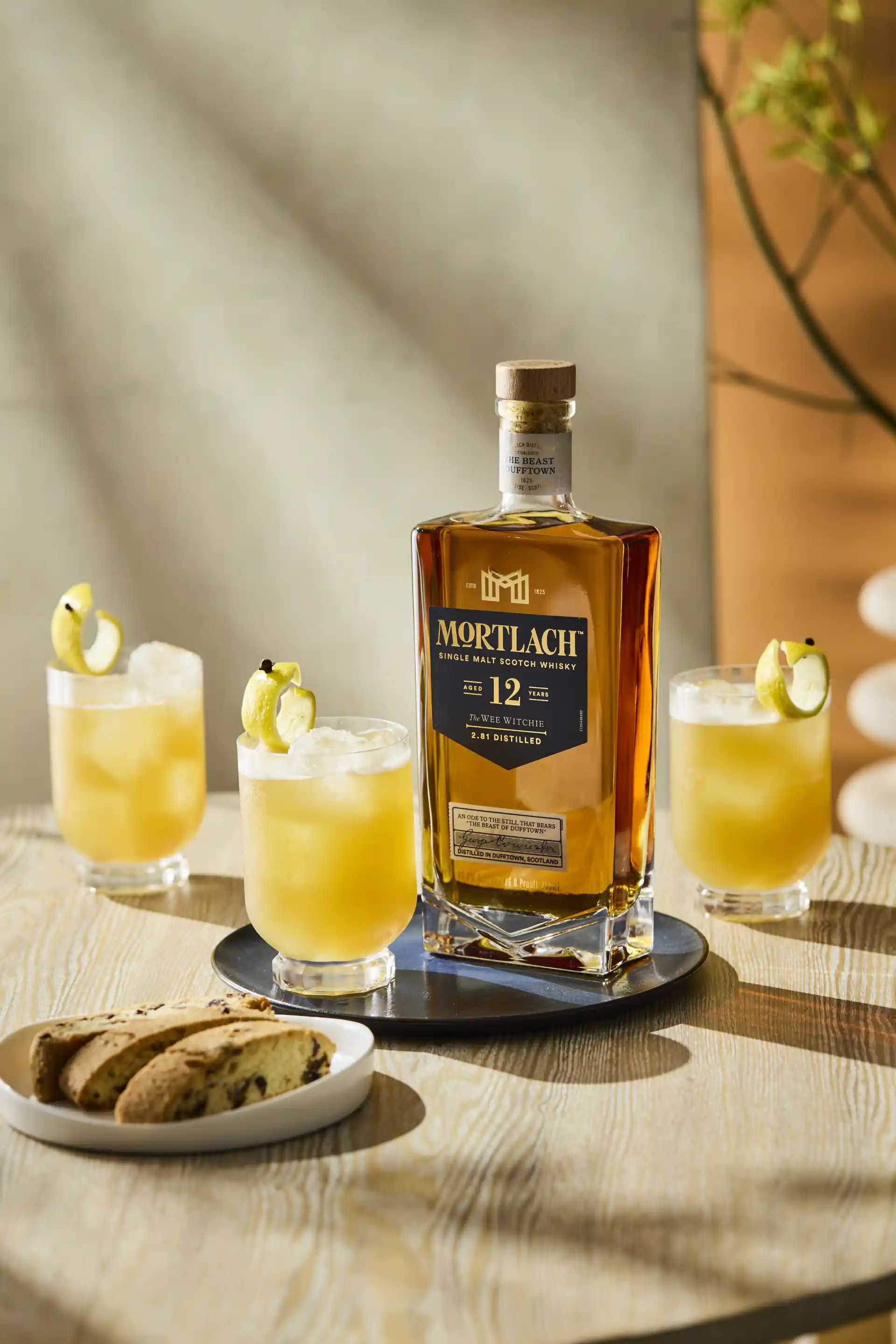 A bottle of Mortlach 12 Year Old stands on a wooden surface with three Pencillin cocktails around it and a plate of cake.