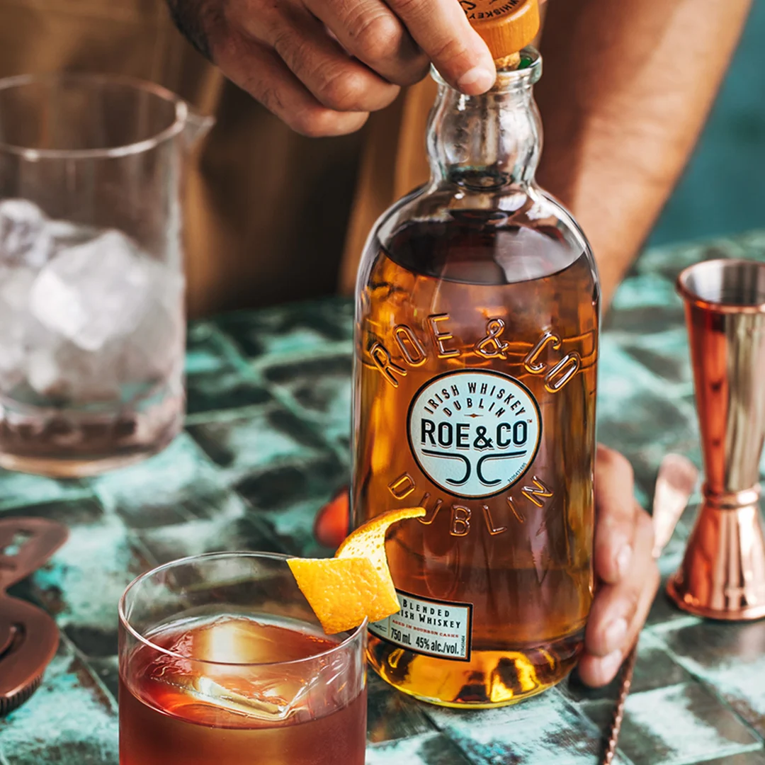 A bottle of Roe &amp; Co whisky sits on a table surrounded by cocktail-making equipment and a cocktail in a rocks glass.