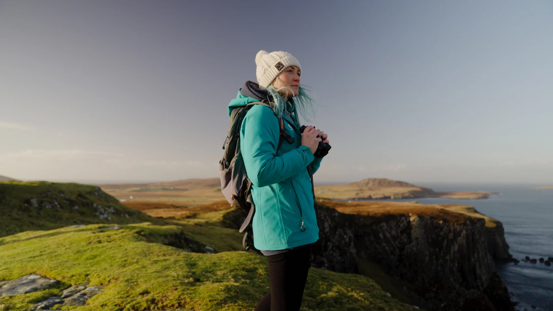 A woman in a blue jacket and white hat stands on a rugged coastline holding a pair of binoculars, looking out to sea.