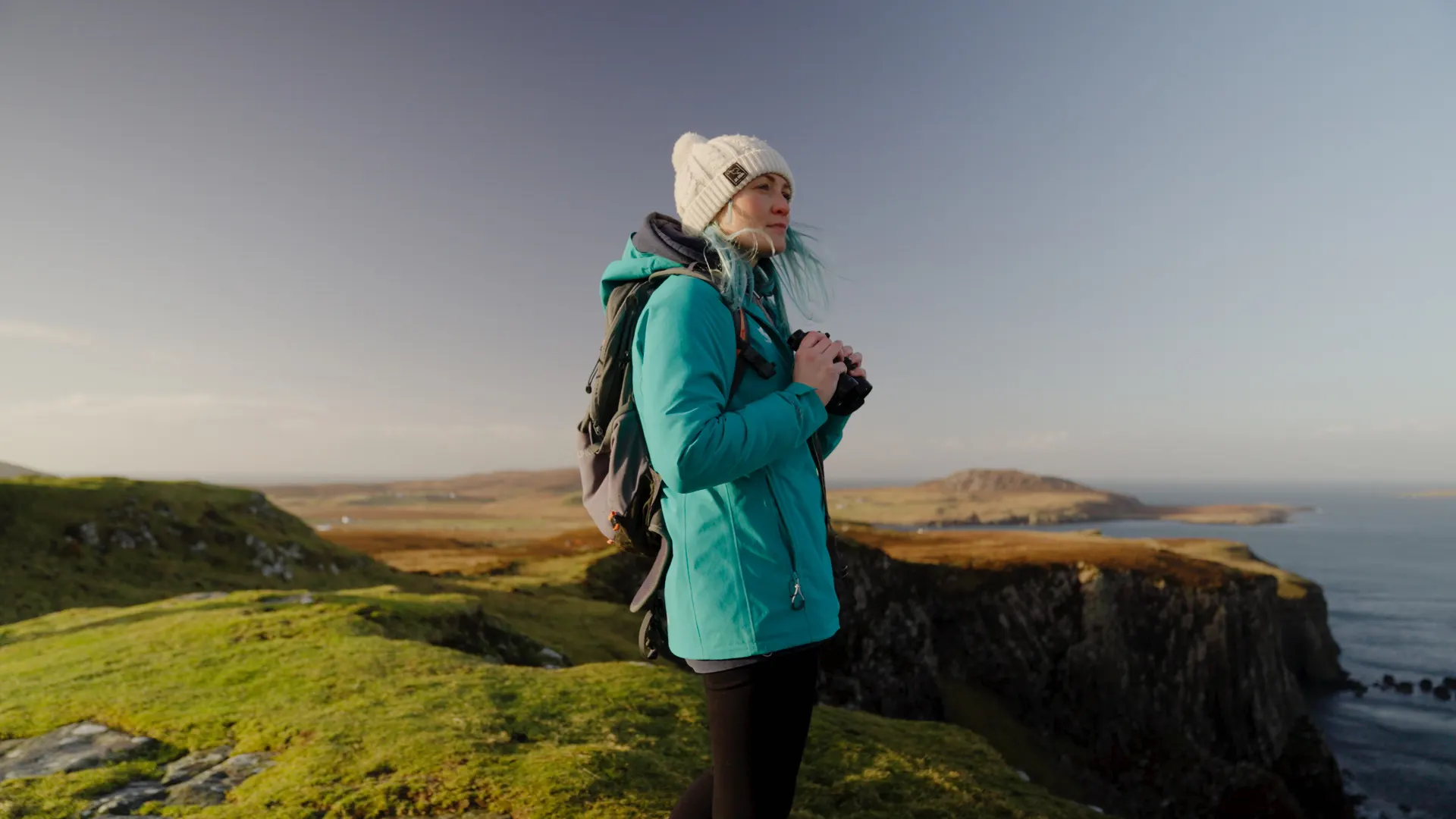 A woman in a blue jacket and white hat stands on a rugged coastline holding a pair of binoculars, looking out to sea.