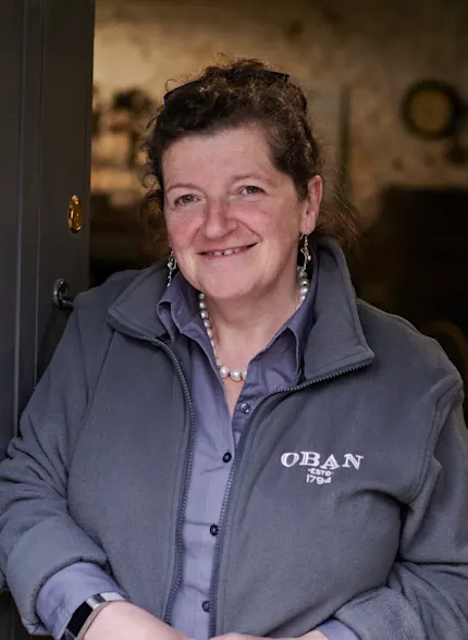 Lesley, eine Reiseleiterin in Oban, steht in einem Lagerraum der Brennerei und trägt eine graue Jacke mit dem Markenzeichen von Oban.