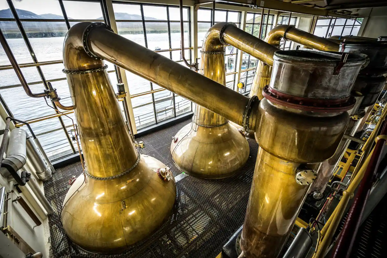 Several metal whisky stills are seen from above, sitting in front of floor to ceiling windows which overlook a body of water.