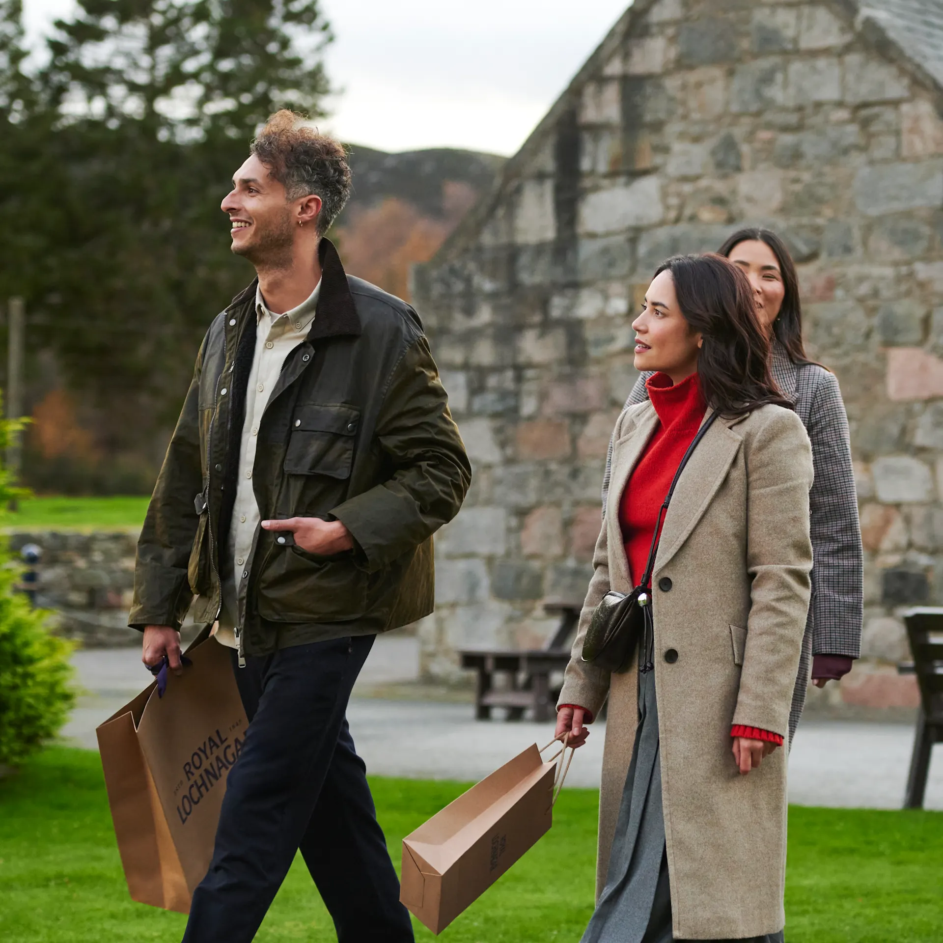 A group of friends leave Royal Lochnagar distillery, walking away from the building and carrying gift bags. They are all smiling.