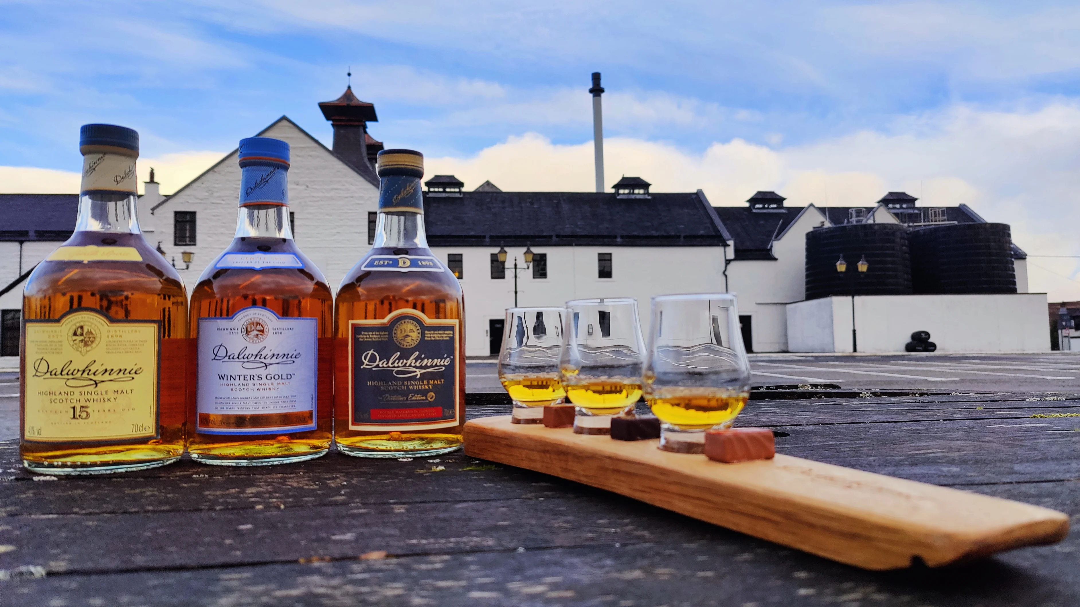 Dalwhinnie Whisky tasting board paired with chocolate and three bottles of Dalwhinnie whisky. Dalwhinnie Distillery in the background.
