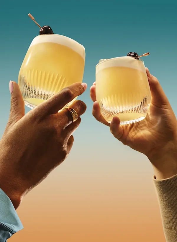 Two hands toasting with whiskey cocktails garnished with cherries against a sunset background