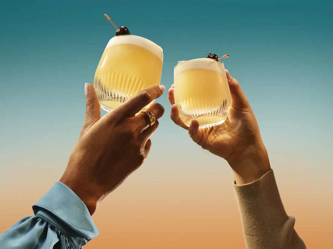 Two hands toasting with whiskey cocktails garnished with cherries against a sunset background
