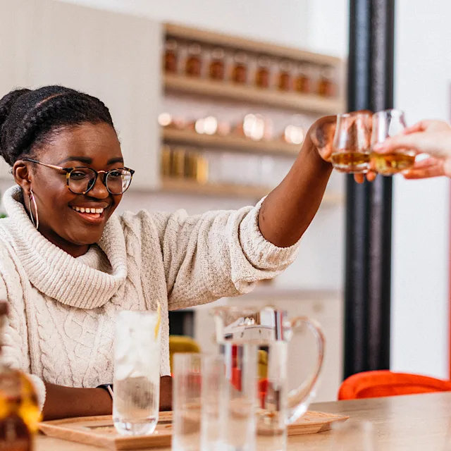 Two women sit either side of a table, each holding a whisky dram above in a ‘cheers’. On the table sits assorted whisky bottles and glasses.
