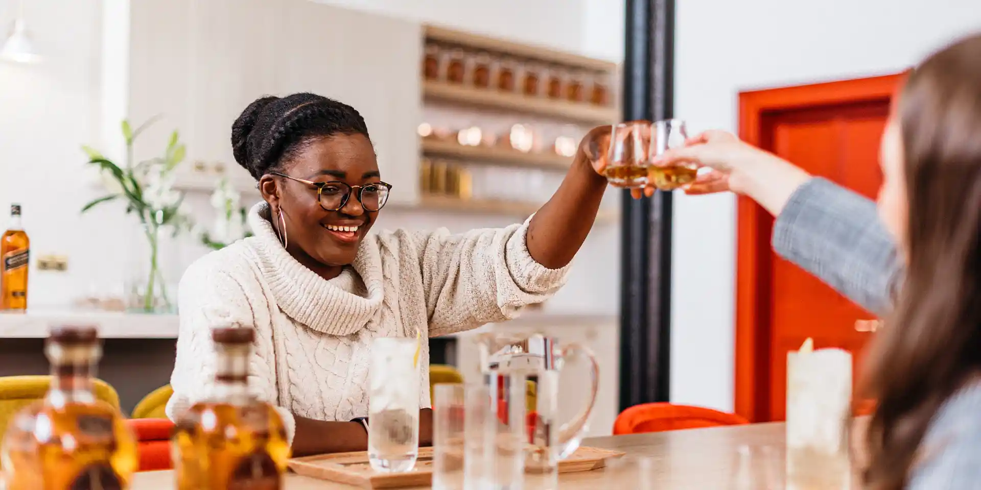 Two women sit either side of a table, each holding a whisky dram above in a ‘cheers’. On the table sits assorted whisky bottles and glasses.