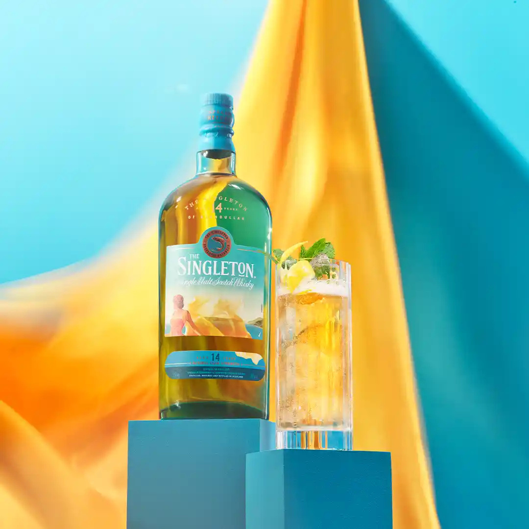 A bottle of The Singleton 14 year old whisky sits on a blue plinth next to a highball glass with a fizzy cocktail in it.