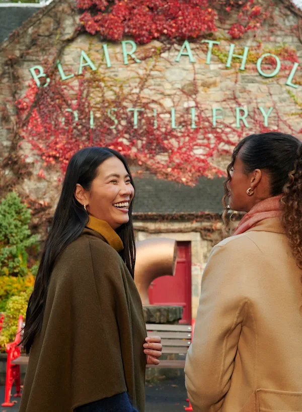 Two female friends stand outside Blair Athol distillery laughing and smiling
