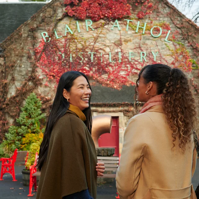 Two female friends stand outside Blair Athol distillery, smiling at each other