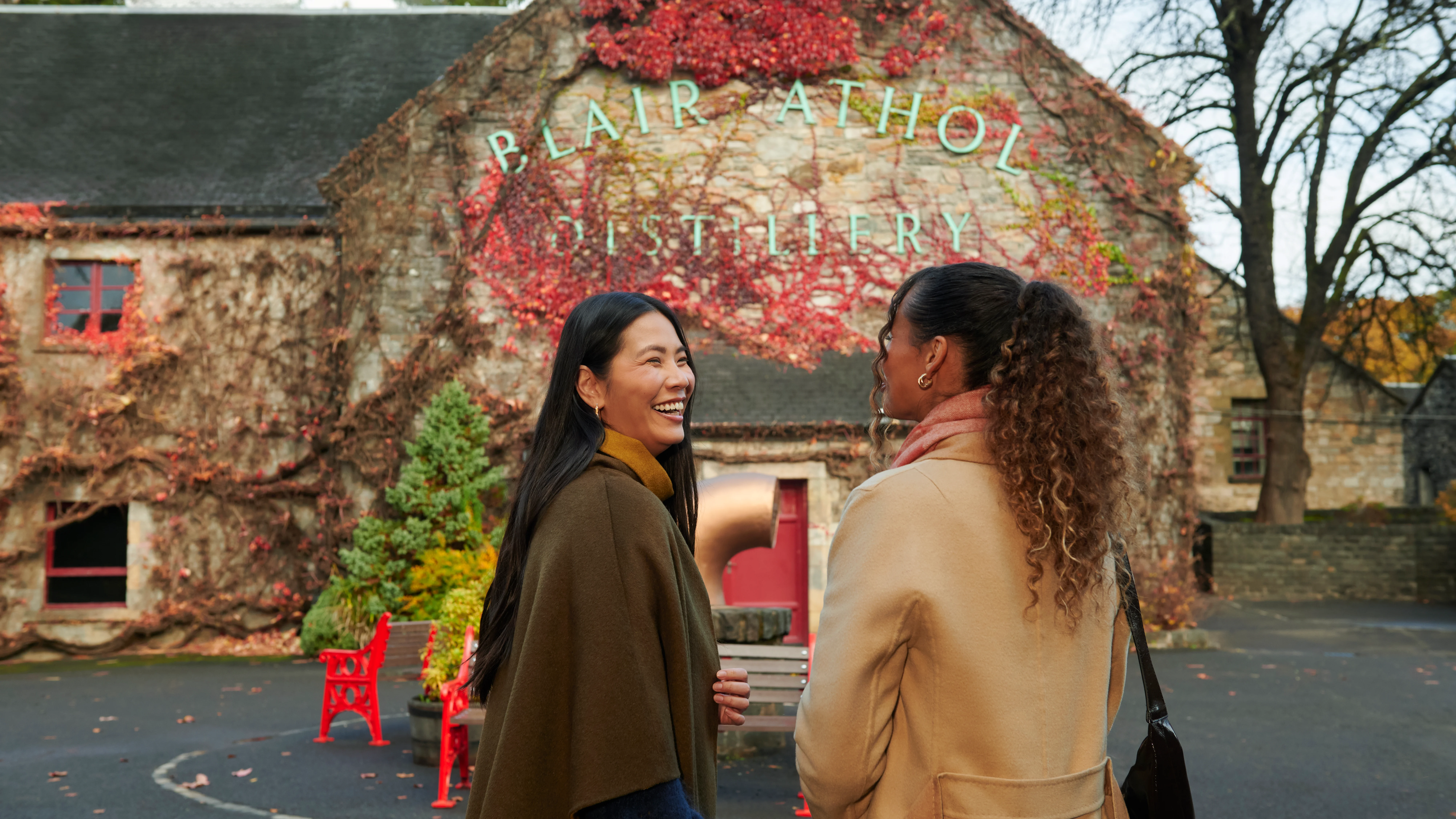 Two female friends stand outside Blair Athol distillery, smiling at each other