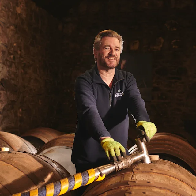 Will Brown, a tour guide from Royal Lochnagar, stands over a whisky barrel holding a hose which is filling the barrel full of whisky. He is smiling.