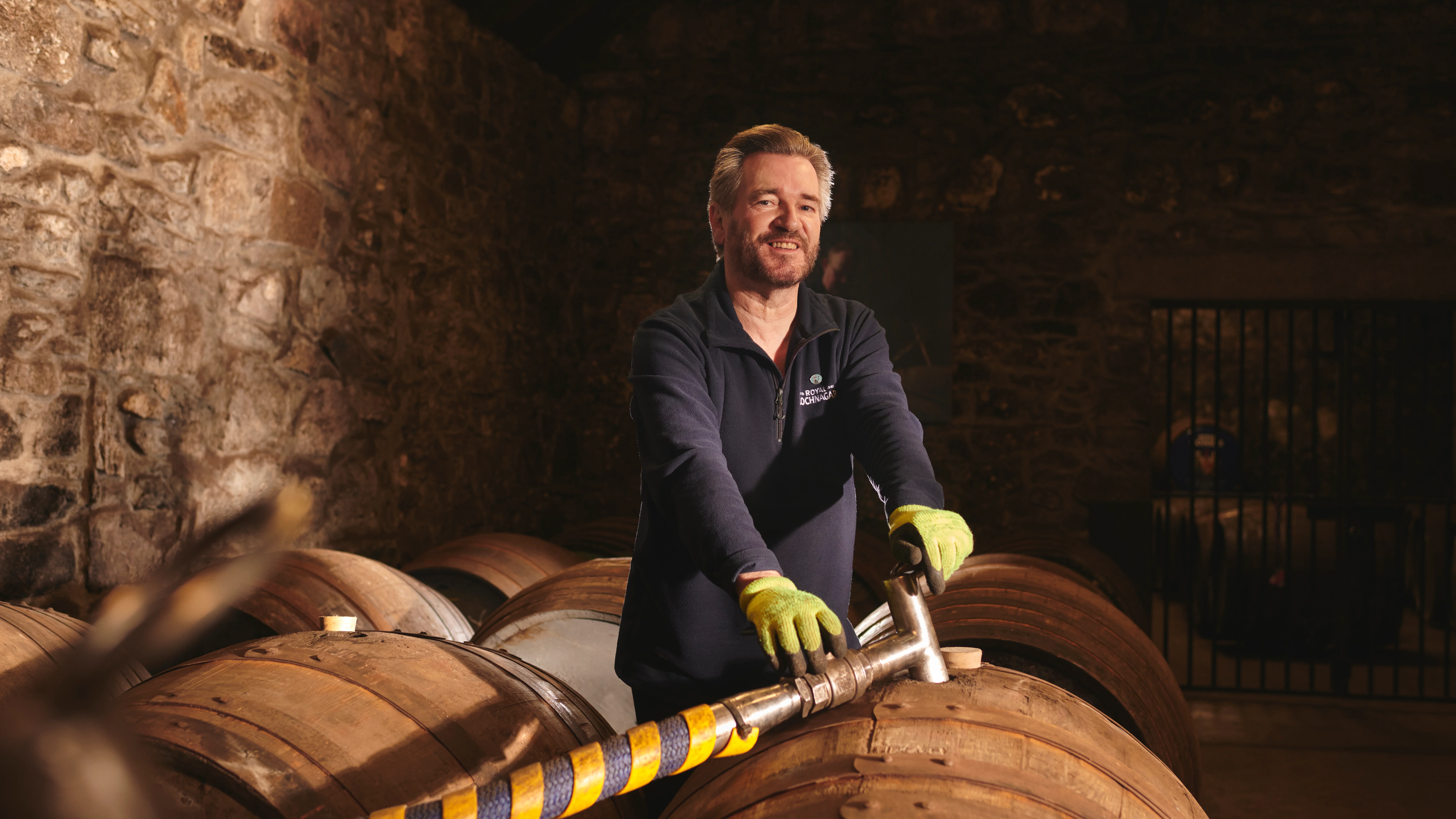 Will Brown, ein Reiseleiter von Royal Lochnagar, steht über einem Whiskyfass und hält einen Schlauch in der Hand, mit dem er das Fass mit Whisky füllt. Er lächelt.