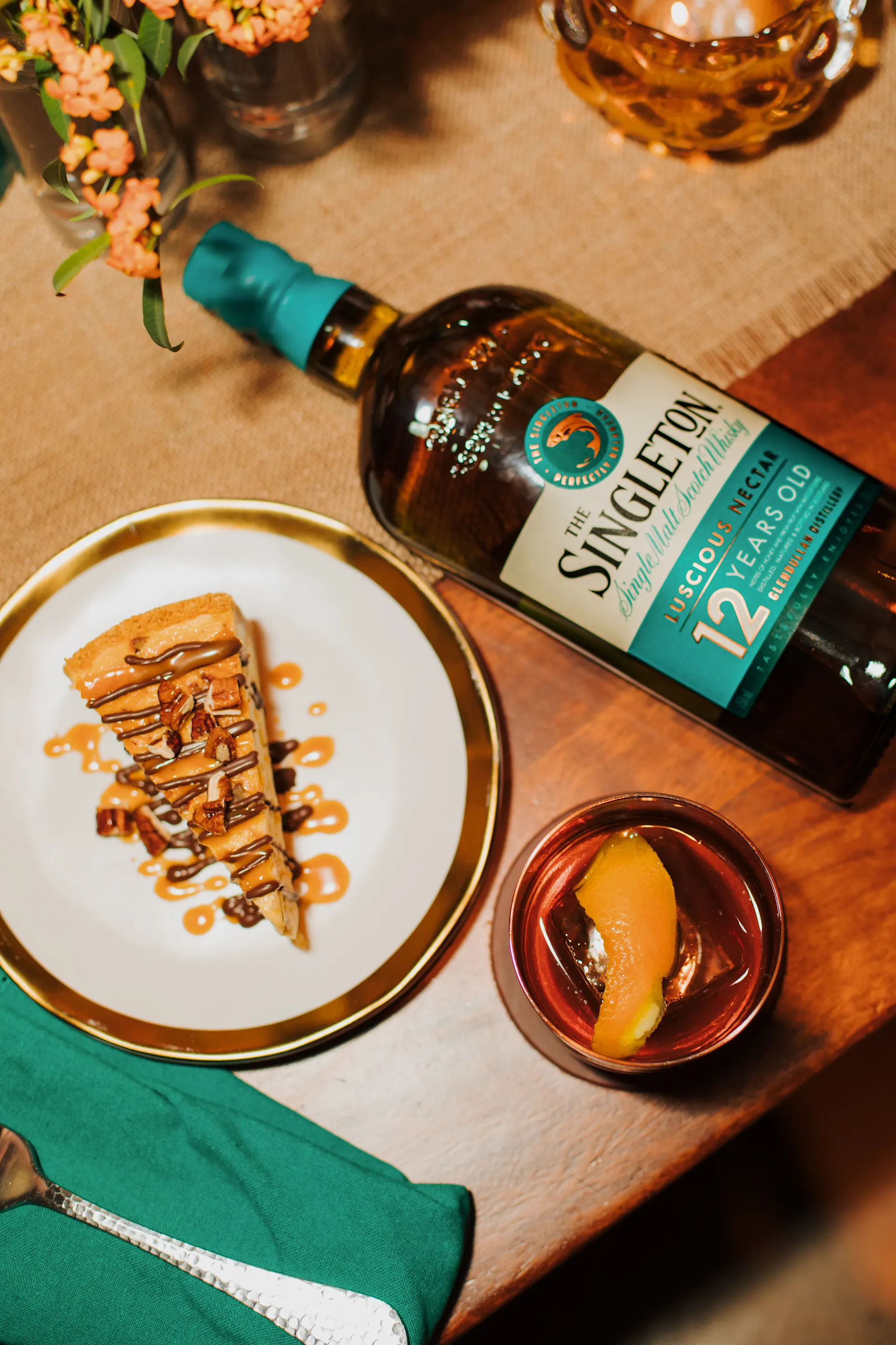 Eine Flasche The Singleton 12 liegt auf dem Rücken auf einem Holztisch, der für eine Mahlzeit hergerichtet ist. Sie liegt neben einem Eisglas mit Whisky-Cocktail und einem Teller mit einem Stück Kuchen.