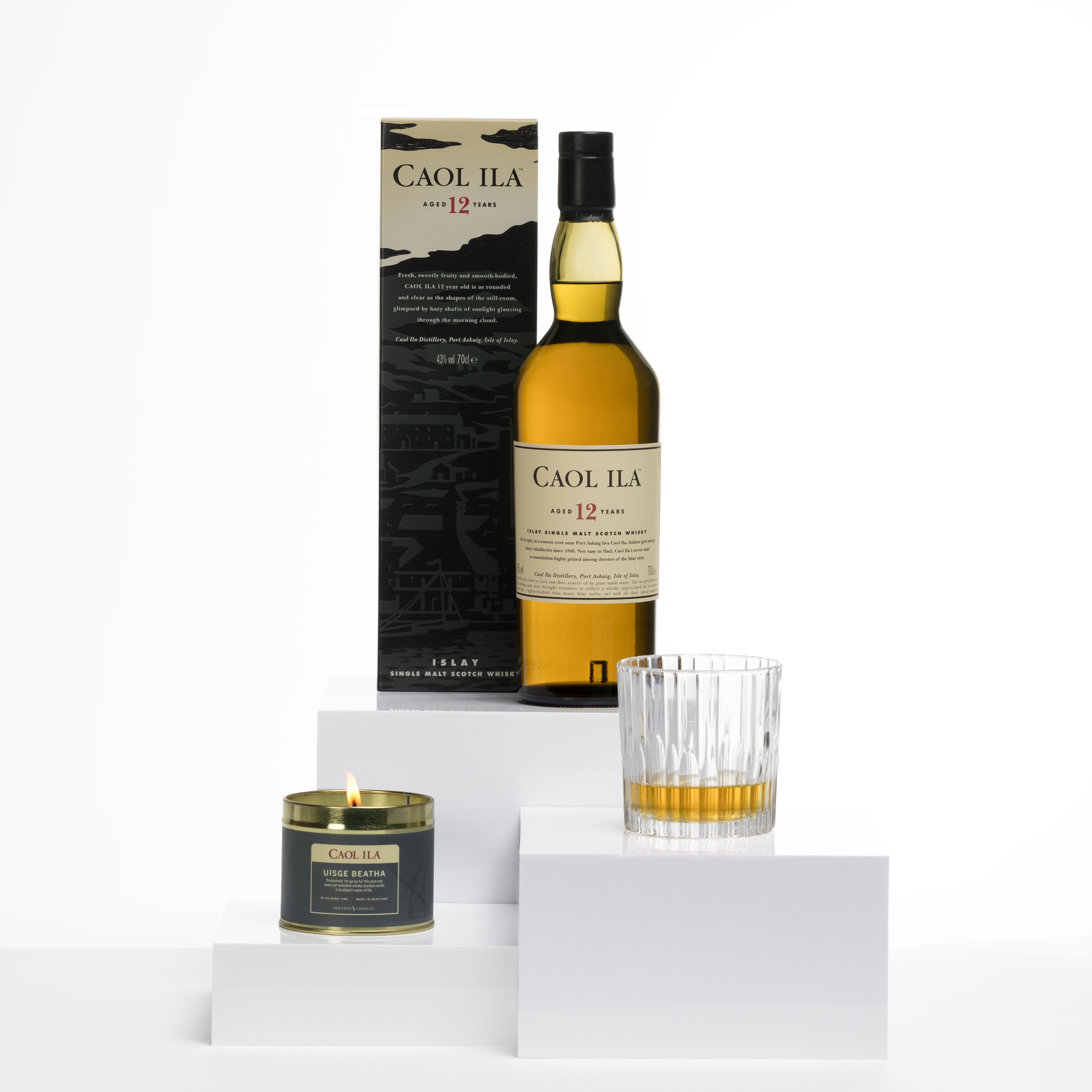 Caol Ila 12 Year Old whiskyfles met een bijpassende doos, een whiskyglas en een brandende whiskygeurkaars op witte displayblokken.