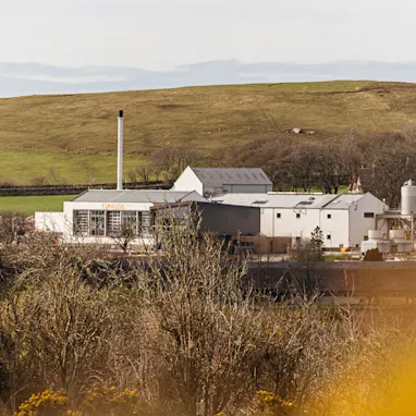 De Clynelish distilleerderij staat tegen een blauwe lucht met witte wolken. Er zijn twee gebouwen - een is wit en op de voorkant staat 'Clynelish'. Het andere is van hout en heeft ramen van vloer tot plafond met een balkon.