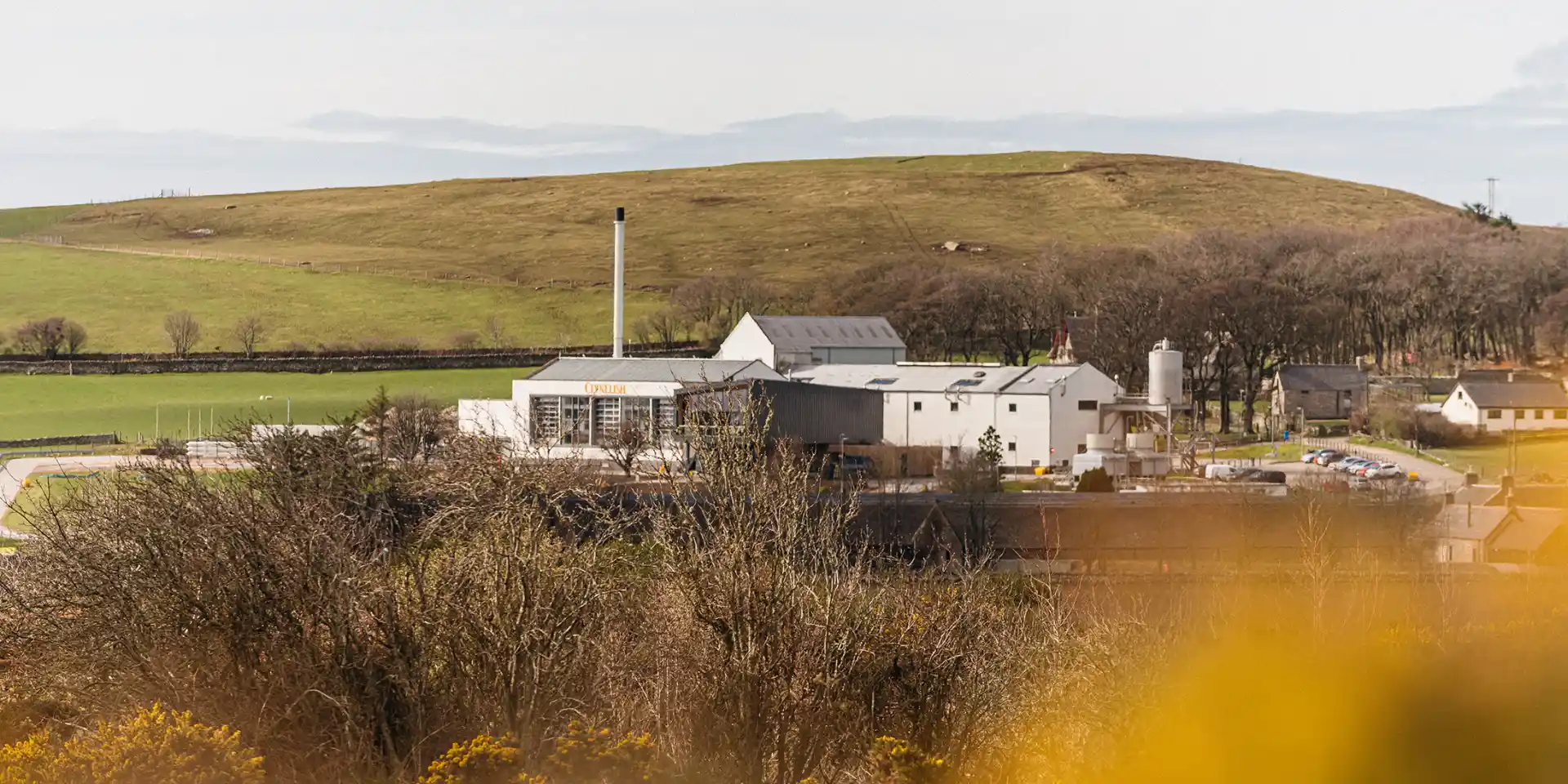 Clynelish distillery stands against a blue sky with white clouds. There are two buildings – one is white and has the words ‘Clynelish’ printed on the front. The other is wooden and features floor to ceiling windows with a balcony.