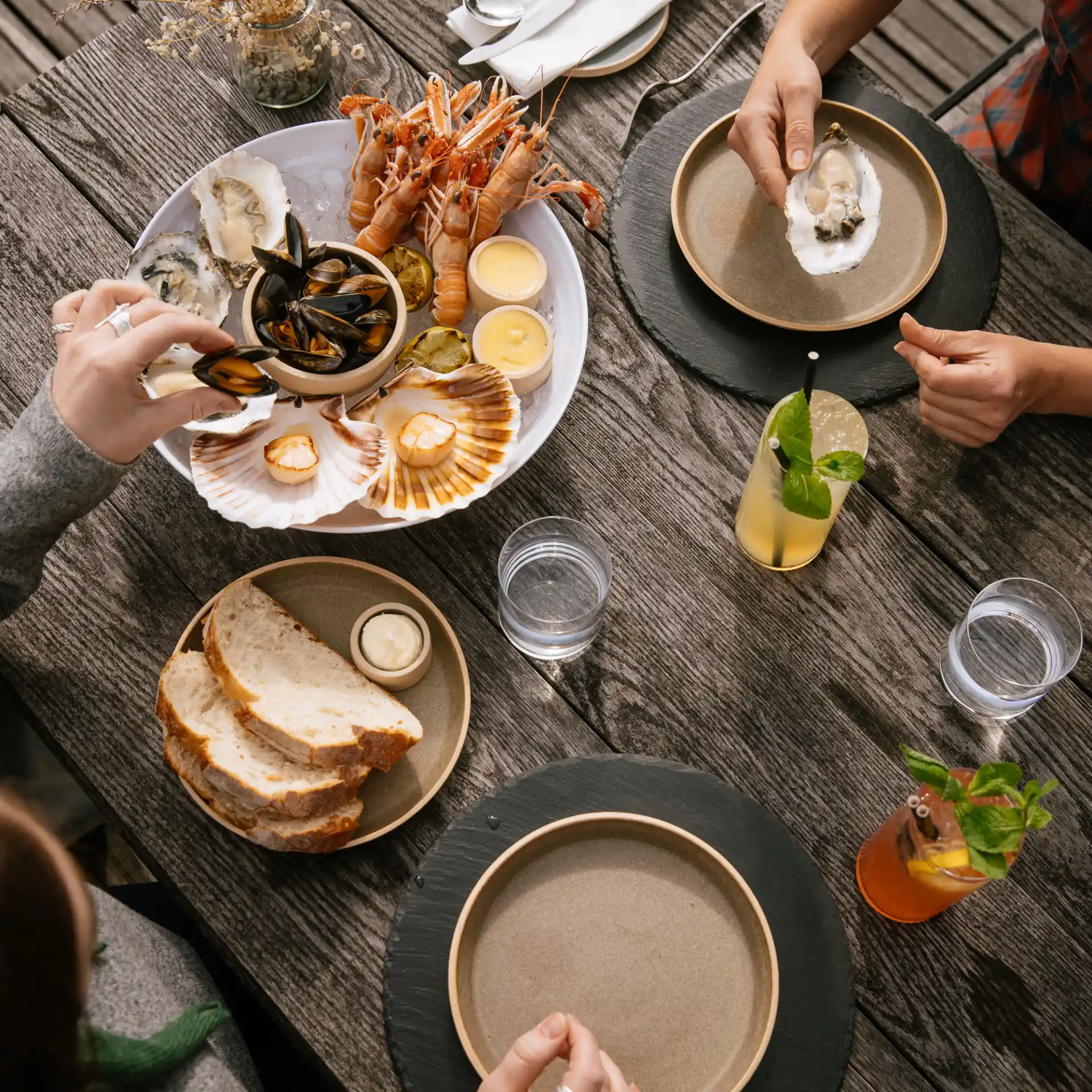 A wooden table is seen from above, with two people sat opposite each other. A bowl of seafood sits on the table, along with a plate of bread and butter and assorted cocktails.