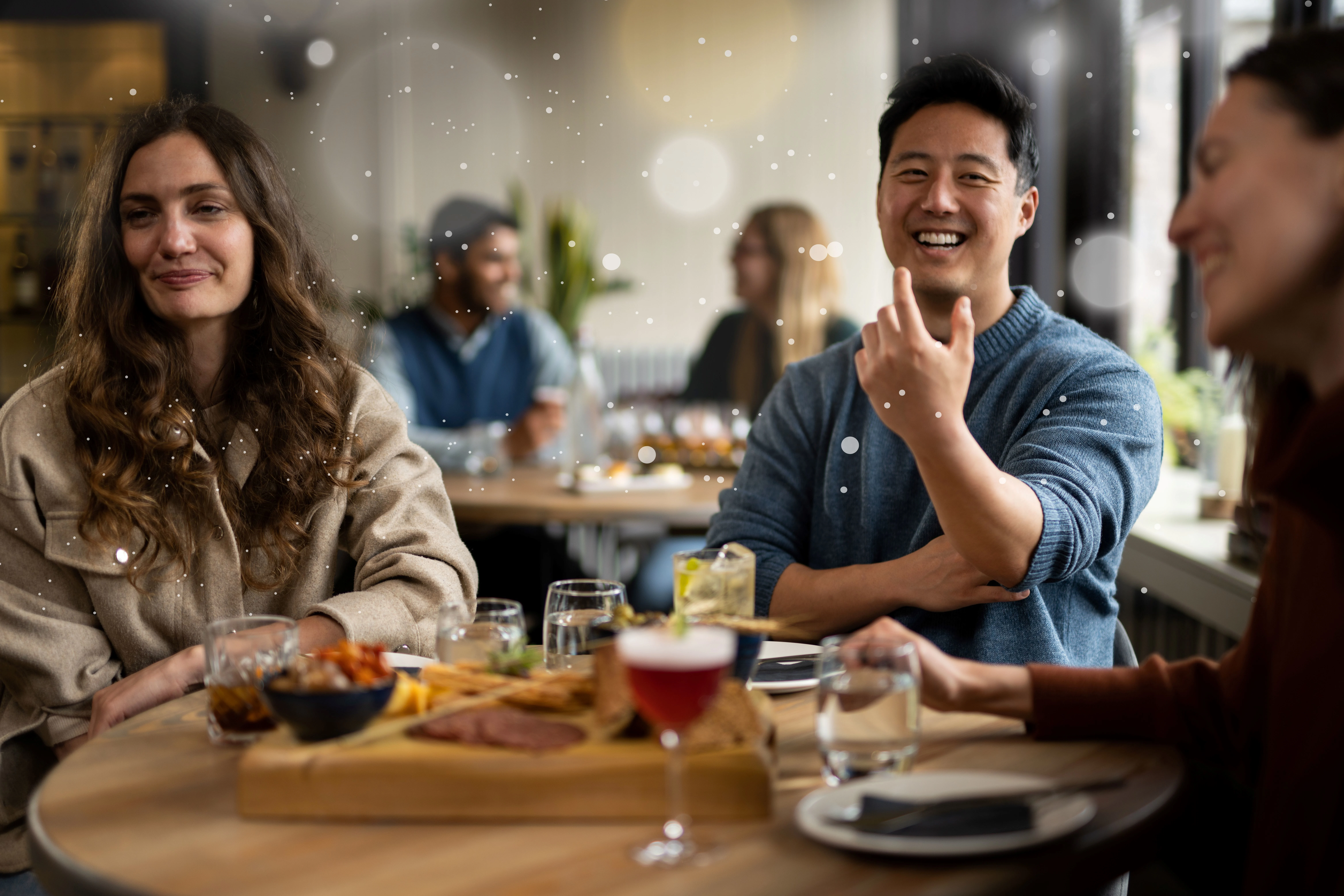 Three friends sitting around a wooden circle table with cocktails and a salame & cheese platter in the middle 