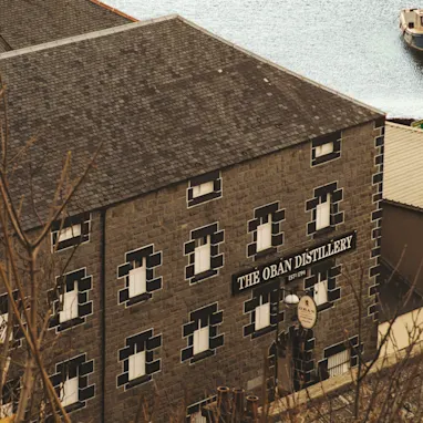 The grey, black and white brick exterior wall of Oban distillery is depicted, with a sign saying The Oban Distillery EST. 1794. The building sits in front of a body of water, a small fishing boat is tethered to a mooring.