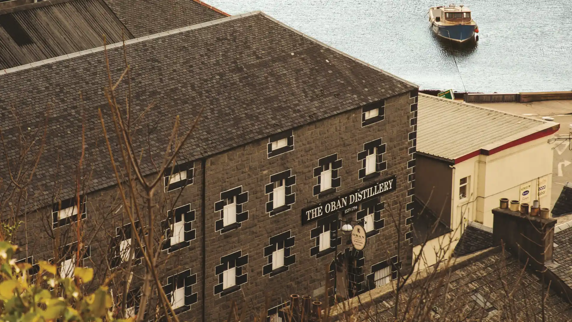 De grijze, zwart-witte bakstenen buitenmuur van de distilleerderij van Oban is afgebeeld, met een bord waarop staat The Oban Distillery EST. 1794. Het gebouw staat voor een waterlichaam, een kleine vissersboot is vastgemaakt aan een meerboei.